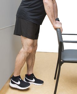 Démonstration de l'exercice 9, on voit que la personne est dos au mur avec une chaise devant elle pour s'appuyer. En levant le pied, son talon touche au mur.