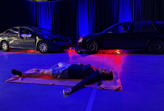Simulation d'un accident de voiture. Une femme blessée est couchée par terre devant deux voitures accidentées.