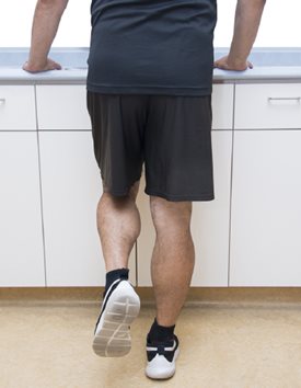 Démonstration de l'exercice 9, la personne a les mains appuyées sur un comptoir et le pied de la jambe non opérée est levé un peu vers l'arrière. De cette façon tout son poids est sur la jambe opérée.